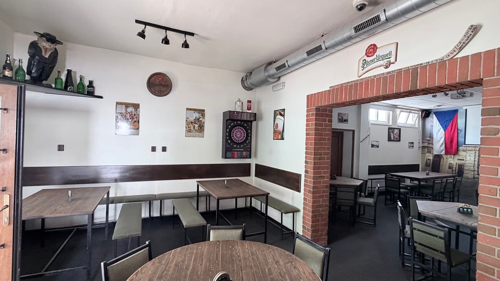Salonek Mlýkárny — šipky, dřevěné stoly a cihlový oblouk do velkého sálu